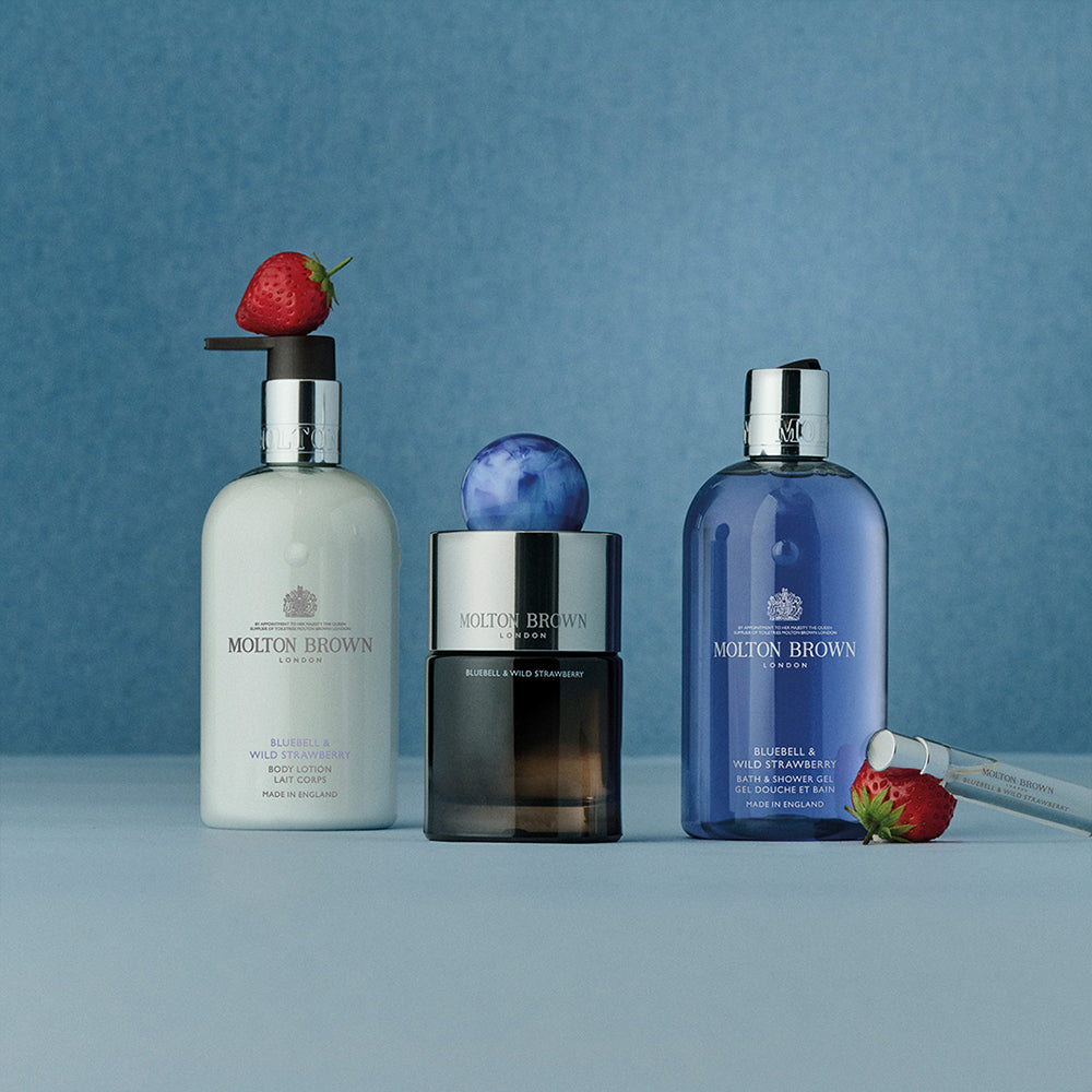 Three MoltonBrown Cyprus products are shown: a hand lotion, hand wash, and Bluebell & Wild Strawberry Eau de Parfum 100ml on a blue backdrop. Wild strawberries surround the bottles, with one on the lotions pump, suggesting a floral-fruity scent.