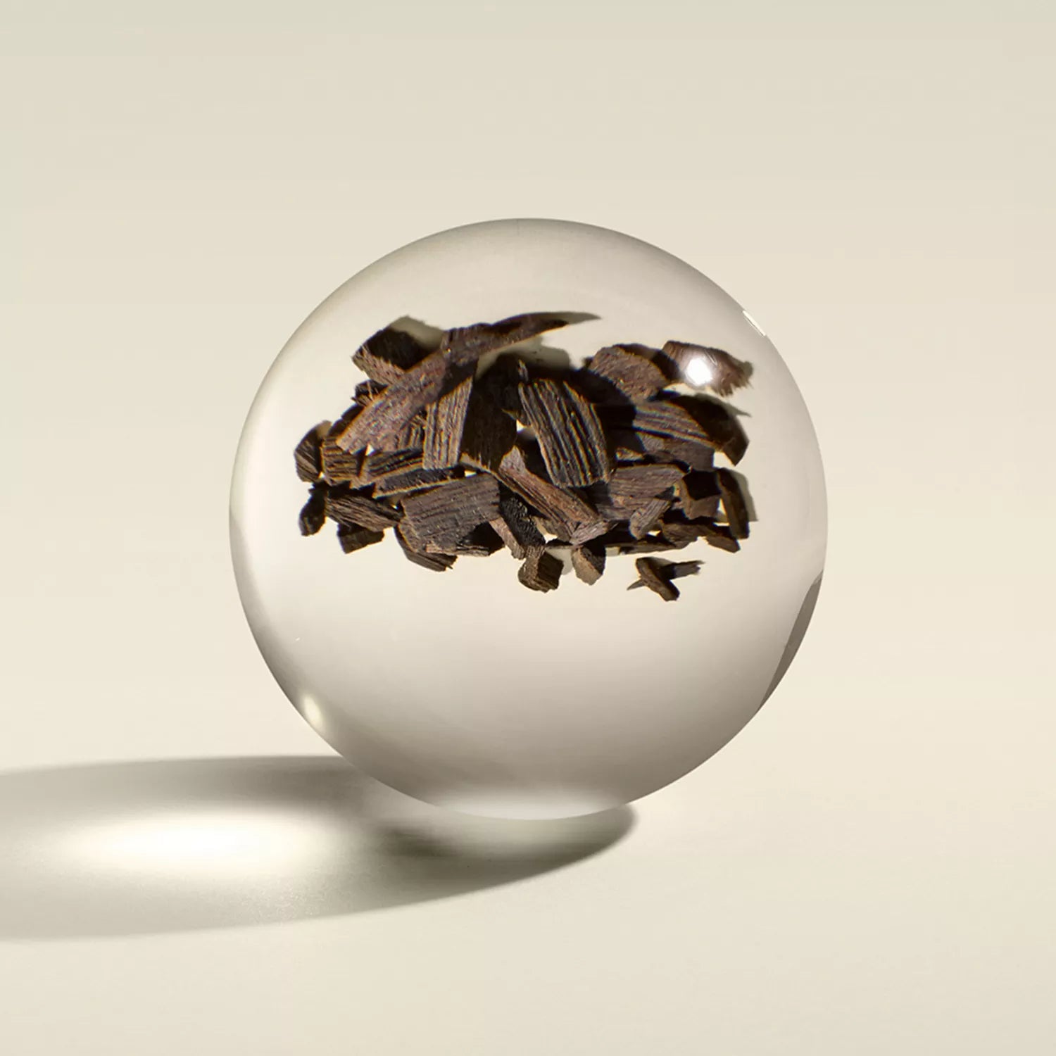 The MoltonBrown Cyprus Mesmerising Oudh Accord & Gold Signature Candle 190g features an opulent clear sphere enclosing jagged dark pieces resembling dried wood chips, evocative of oudh, casting a soft shadow on a light beige surface with the contents appearing suspended inside.