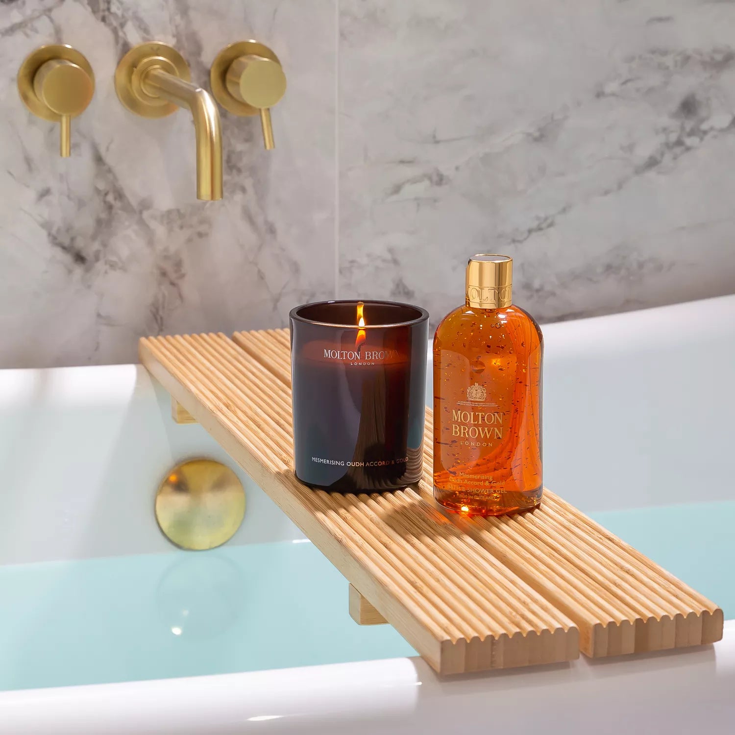 A bathtub with a wooden tray holds a Mesmerising Oudh Accord & Gold Signature Candle by MoltonBrown Cyprus and an orange Molton Brown bottle. The water-filled tub, adorned by gold faucets on a marble wall, creates a luxurious oasis.