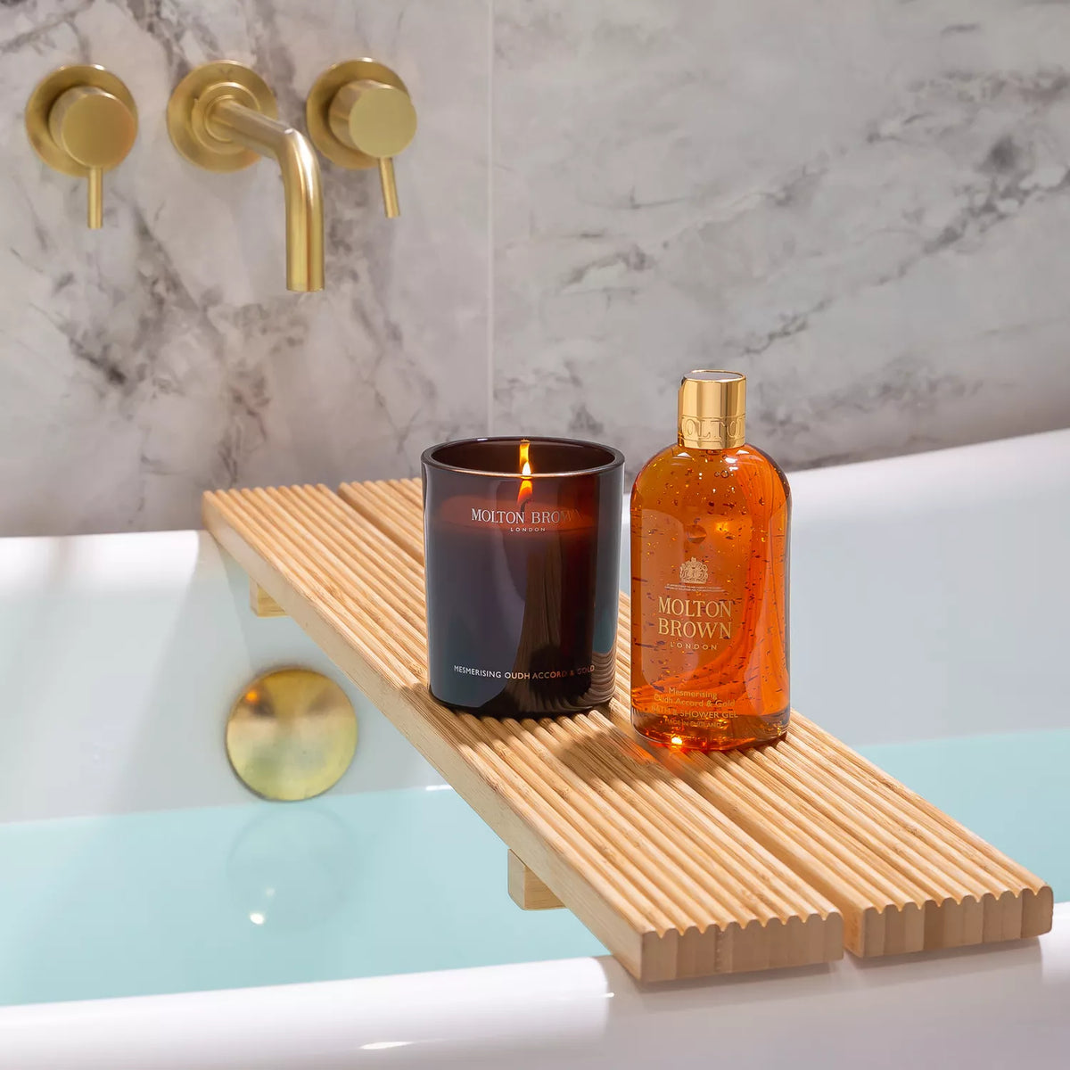 A bathtub with a wooden tray holds a Mesmerising Oudh Accord & Gold Signature Candle by MoltonBrown Cyprus and an orange Molton Brown bottle. The water-filled tub, adorned by gold faucets on a marble wall, creates a luxurious oasis.