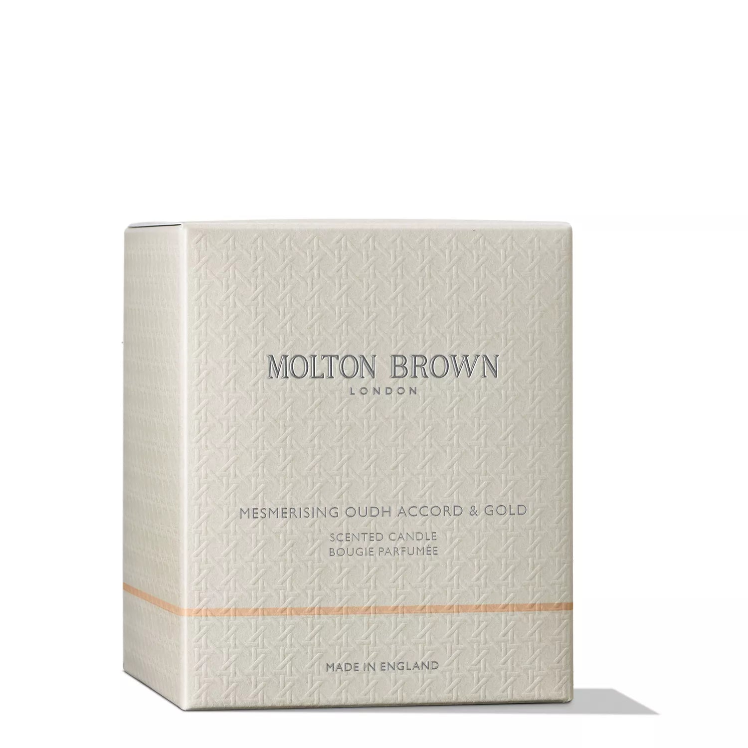 A beige embossed MoltonBrown Cyprus box, housing the Mesmerising Oudh Accord & Gold Signature Candle 190g with hints of cinnamon, displays Molton Brown London, Mesmerising Oudh Accord & Gold, Scented Candle, Bougie Parfumée on the front. An orange line graces the bottom. Made in England for luxury candle enthusiasts.