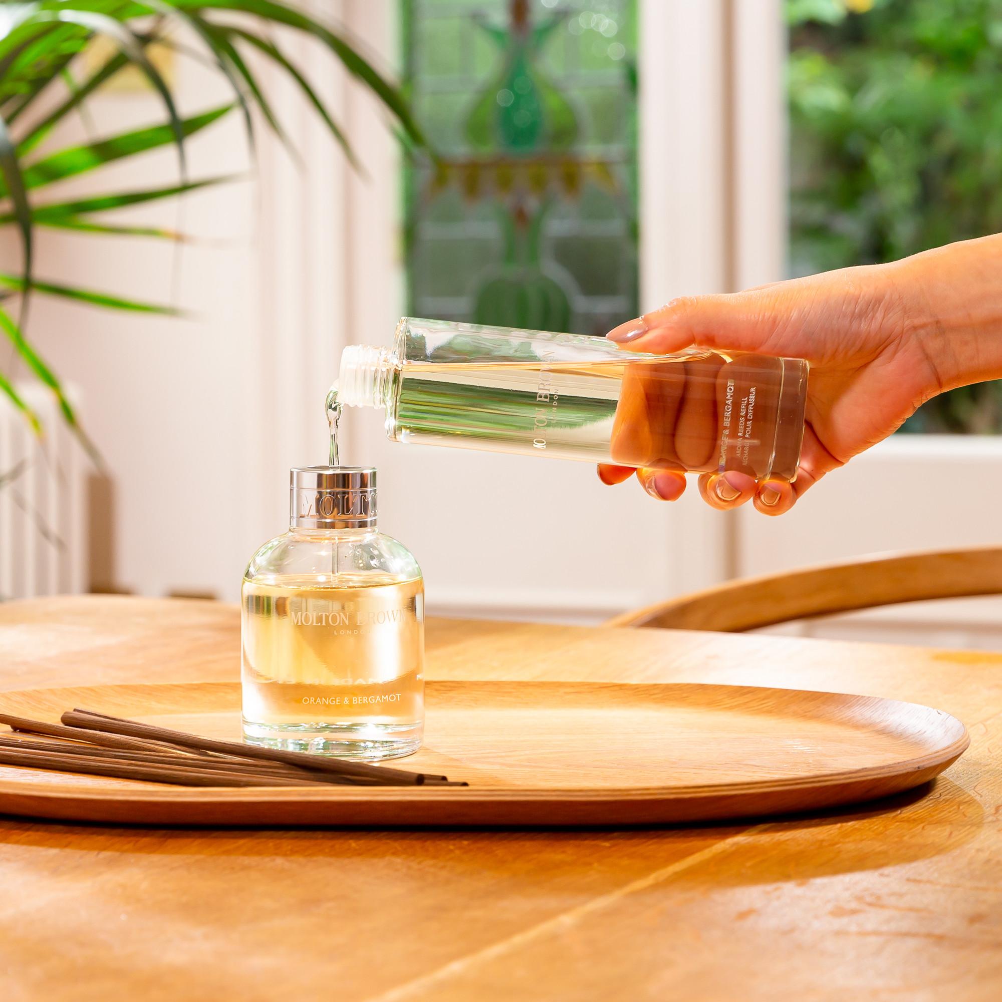 A hand pours liquid into a clear fragrance diffuser bottle on a wooden tray, surrounded by scattered reed sticks. Sunlight highlights a green plant in the background, enhancing the invigorating scent of Orange & Bergamot from MoltonBrown Cyprus's Aroma Reeds Refill 150ml.