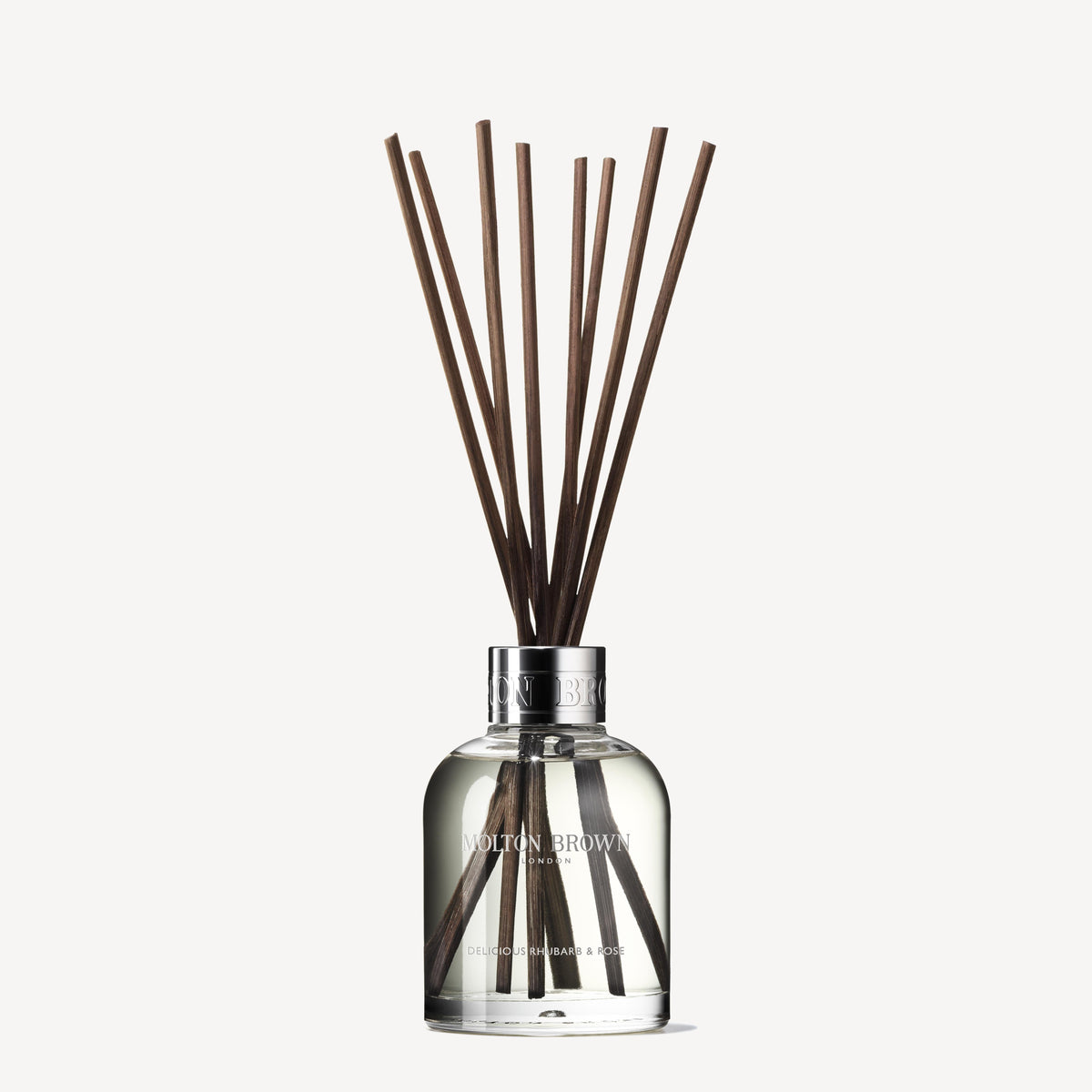The Delicious Rhubarb & Rose Aroma Reeds 150ml from MoltonBrown Cyprus comes in a clear glass bottle, showcasing several long, dark brown reeds that extend upward. The visible liquid emits enticing exotic rhubarb and rose scents. A silver cap adorned with the brand name completes this sophisticated accessory, promising a luxurious aroma.