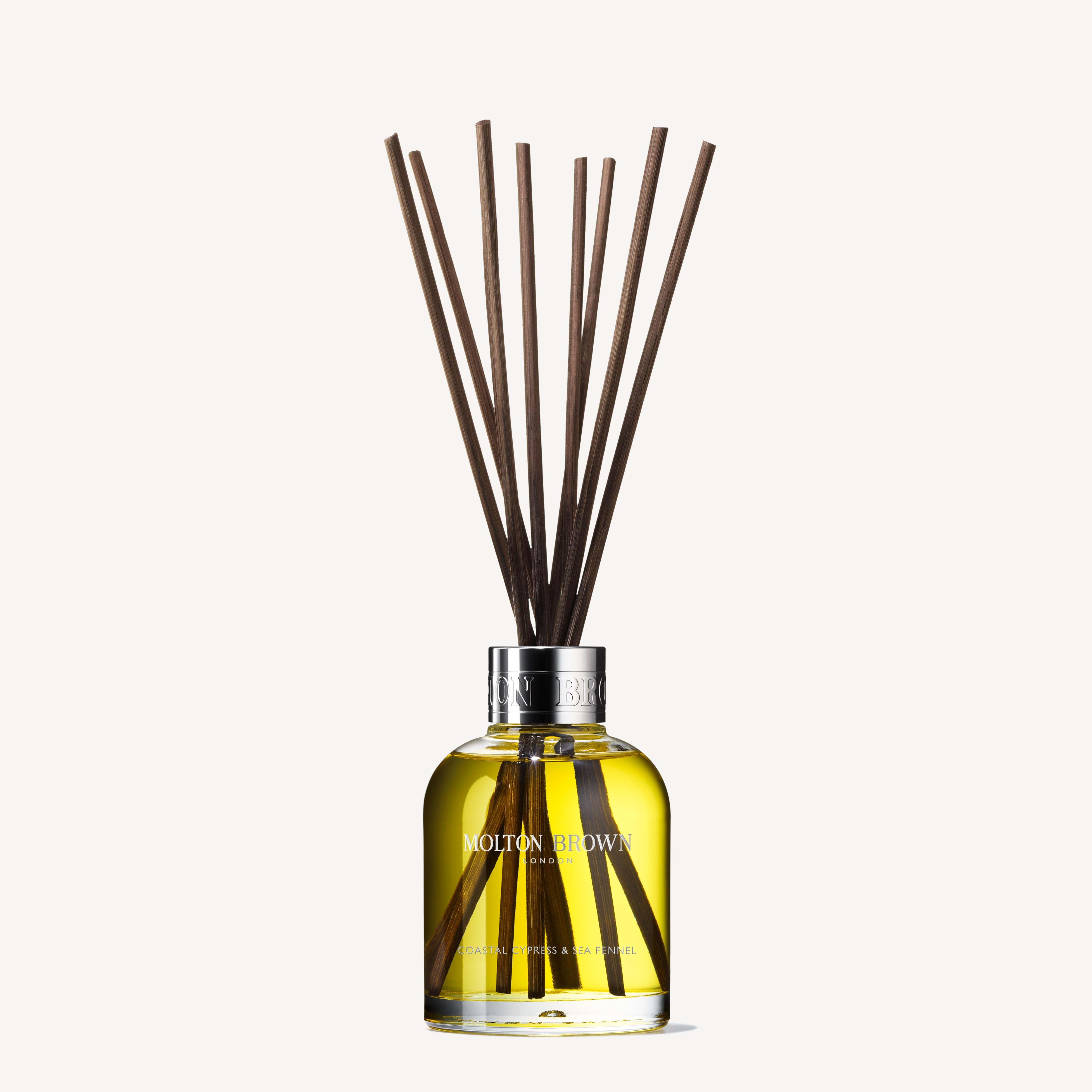 A transparent glass bottle, elegantly branded MoltonBrown Cyprus, contains a fragrant yellow liquid. From its top, dark brown aroma reeds rise gracefully, releasing the scent of Coastal Cypress & Sea Fennel into the air. This sophisticated arrangement is set against a simple white background.