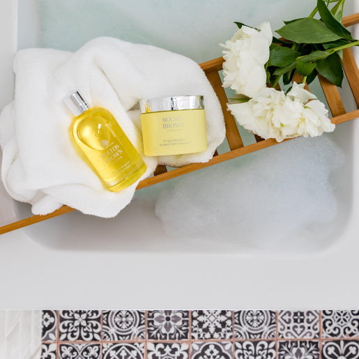 A luxurious bath scene with a wooden tray holding crisp white towels, the Molton Brown Cyprus Orange & Bergamot Radiant Body Polisher, a jar of cream, and pristine white peonies over a bubble-filled tub. The floor features intricately designed black and white patterned tiles.