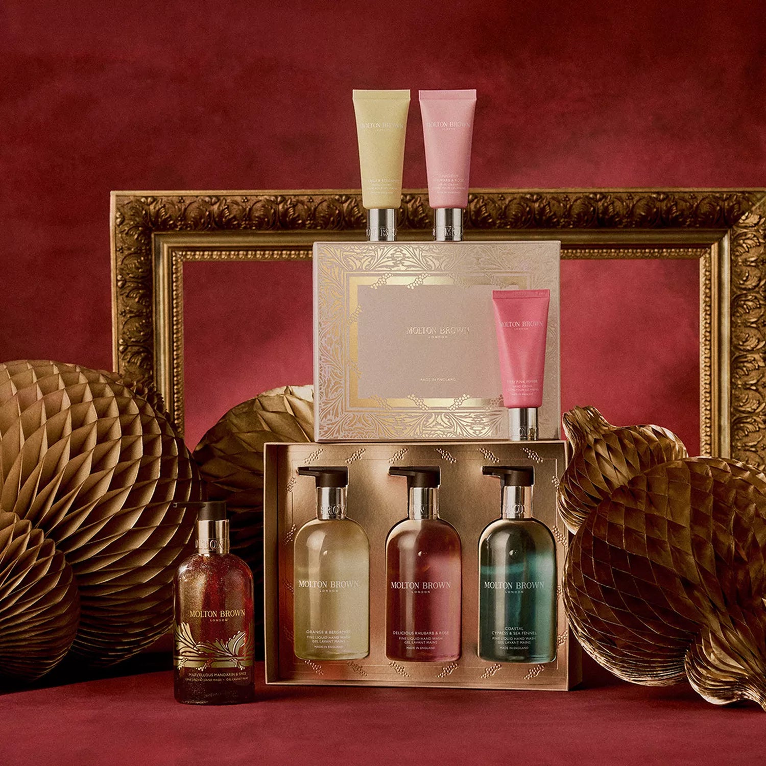 The Floral & Fresh Hand Care Collection by MoltonBrown Cyprus features Fine Liquid Hand Wash, elegantly packaged, set against a rich red backdrop with gold frames and honeycomb décor, offering long-lasting fragrance in every bottle and tube.