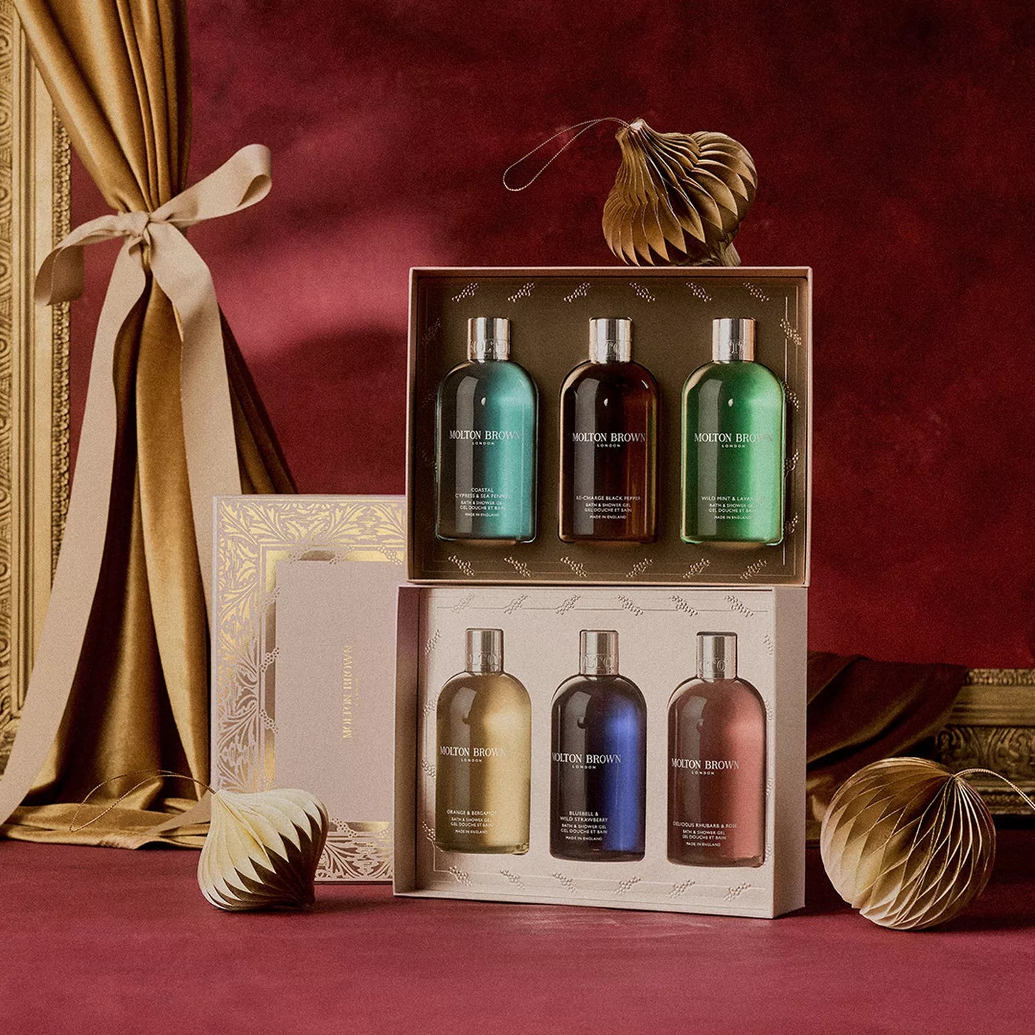 Two Woody & Fresh Body Care Gift Sets by MoltonBrown Cyprus, each with four colorful bath & shower gels, are showcased on a rich red surface with gold accents and a ribbon—ideal for anyone who loves restorative body wash.