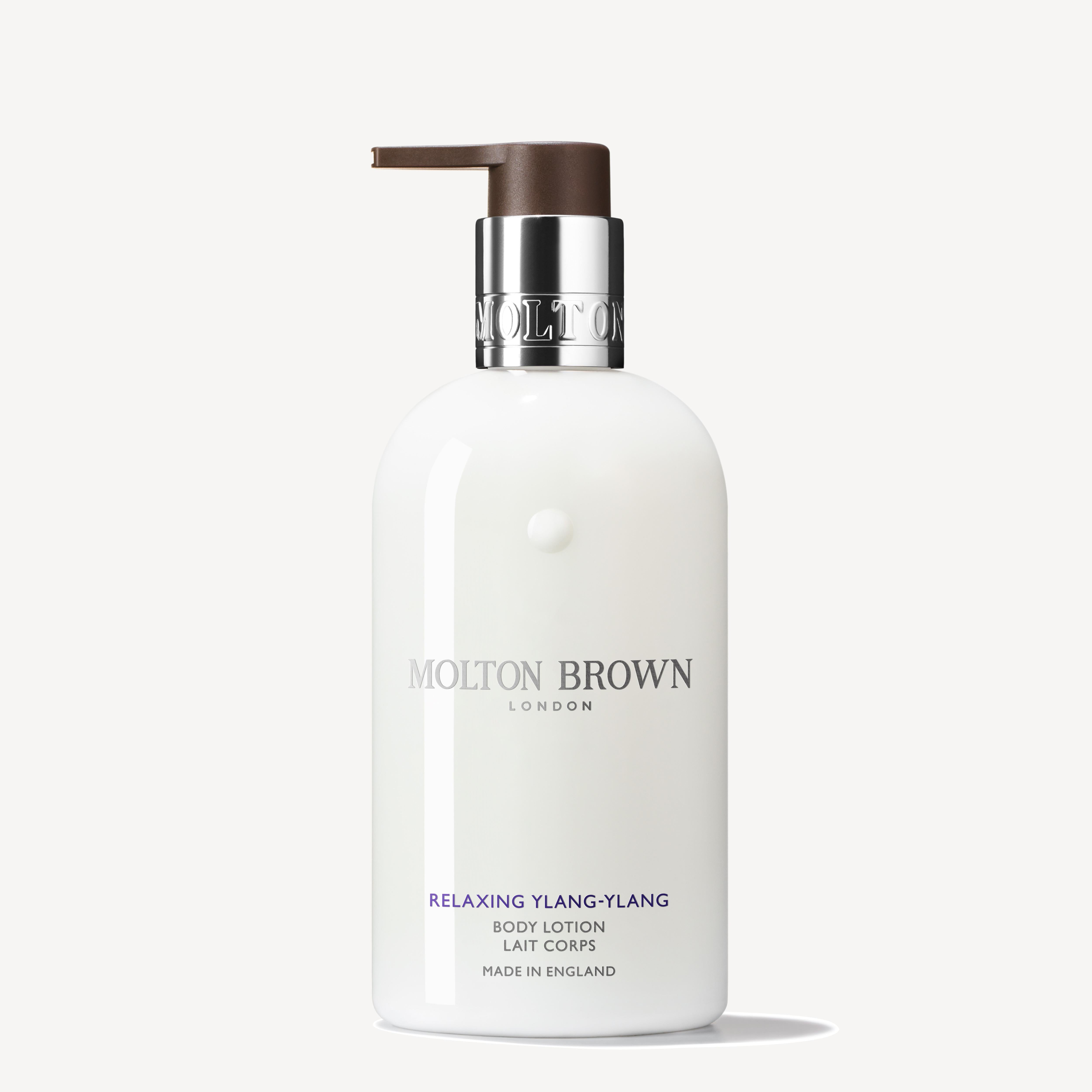 A white bottle of MoltonBrown Cyprus Relaxing Ylang-Ylang Body Lotion, infused with cardamom, comes with a brown pump dispenser. The label highlights the brand logo and product information, showcasing its English craftsmanship. The 300ml moisturizing body lotion is displayed against a simple light background.