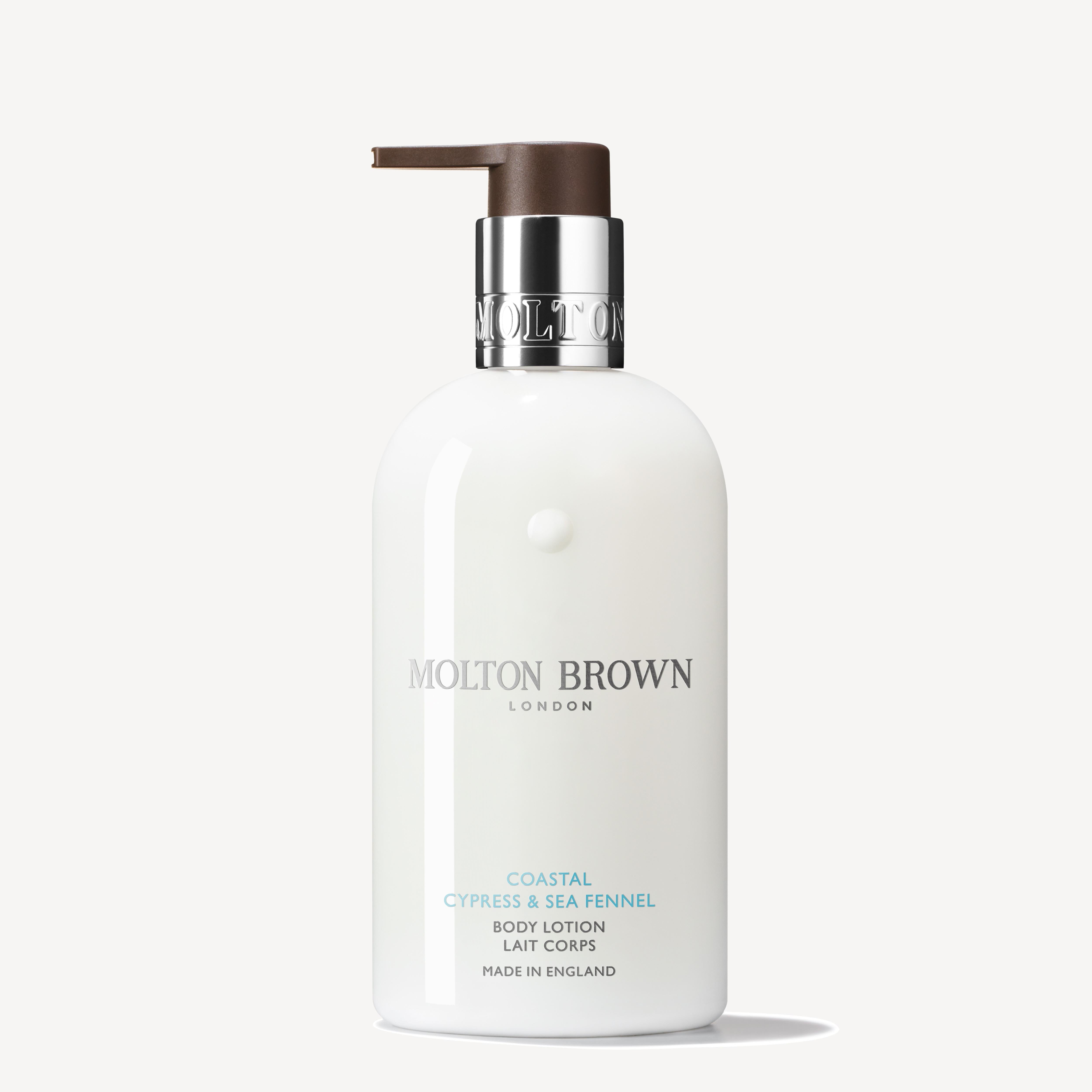 A 300ml bottle of MoltonBrown Cyprus Coastal Cypress & Sea Fennel Body Lotion, featuring a stylish white design with a silver and brown pump dispenser, elegantly displays the brand's logo and text in silver and blue while hydrating the skin beautifully.