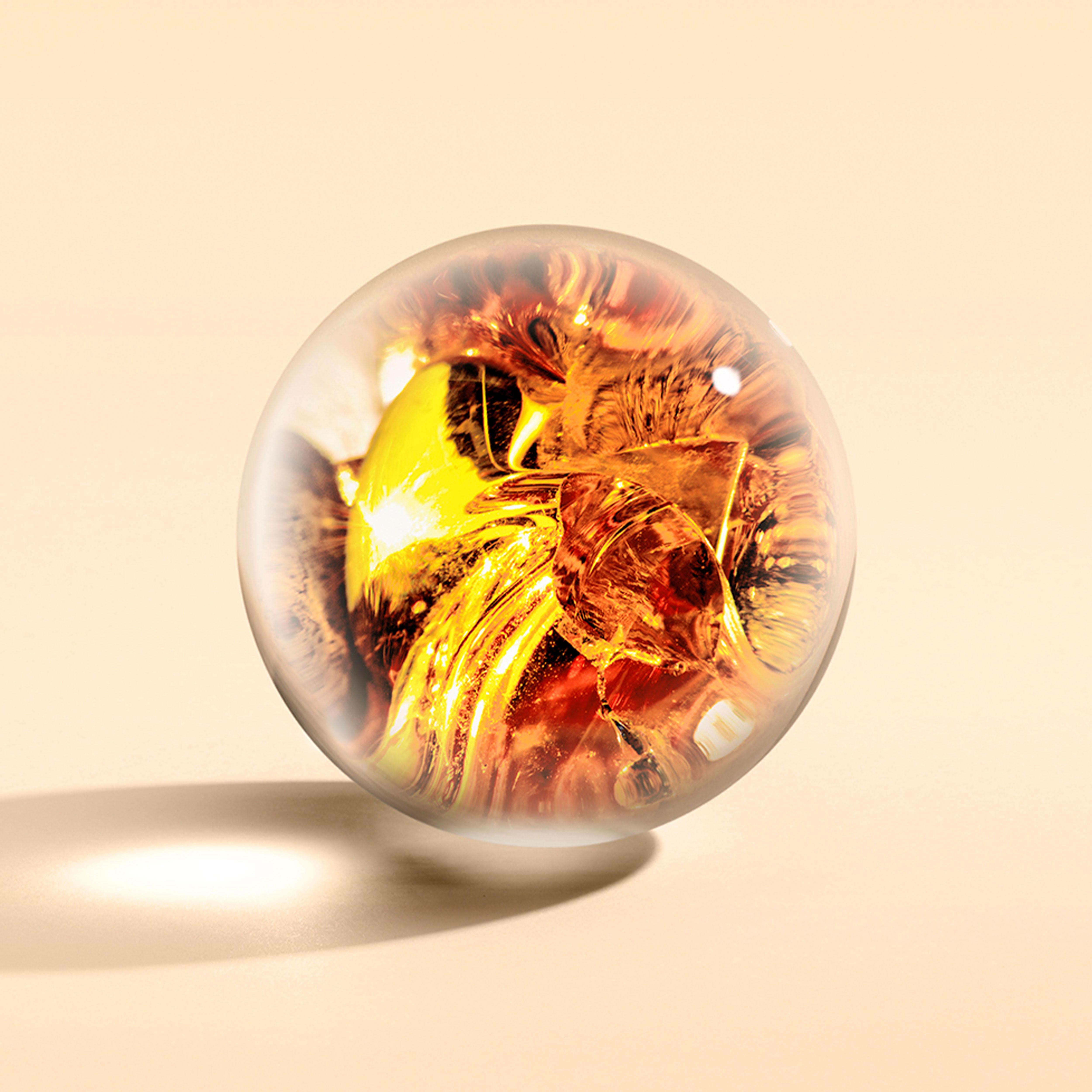 A bottle of Neon Amber Body Lotion by MoltonBrown Cyprus rests on a light surface, with its clear glass sphere packaging featuring molten golden-yellow swirls that resemble flowing liquid or flames infused with the essence of mandarin oil. This dynamic design creates a warm ambiance while light reflections add depth to its spherical form.