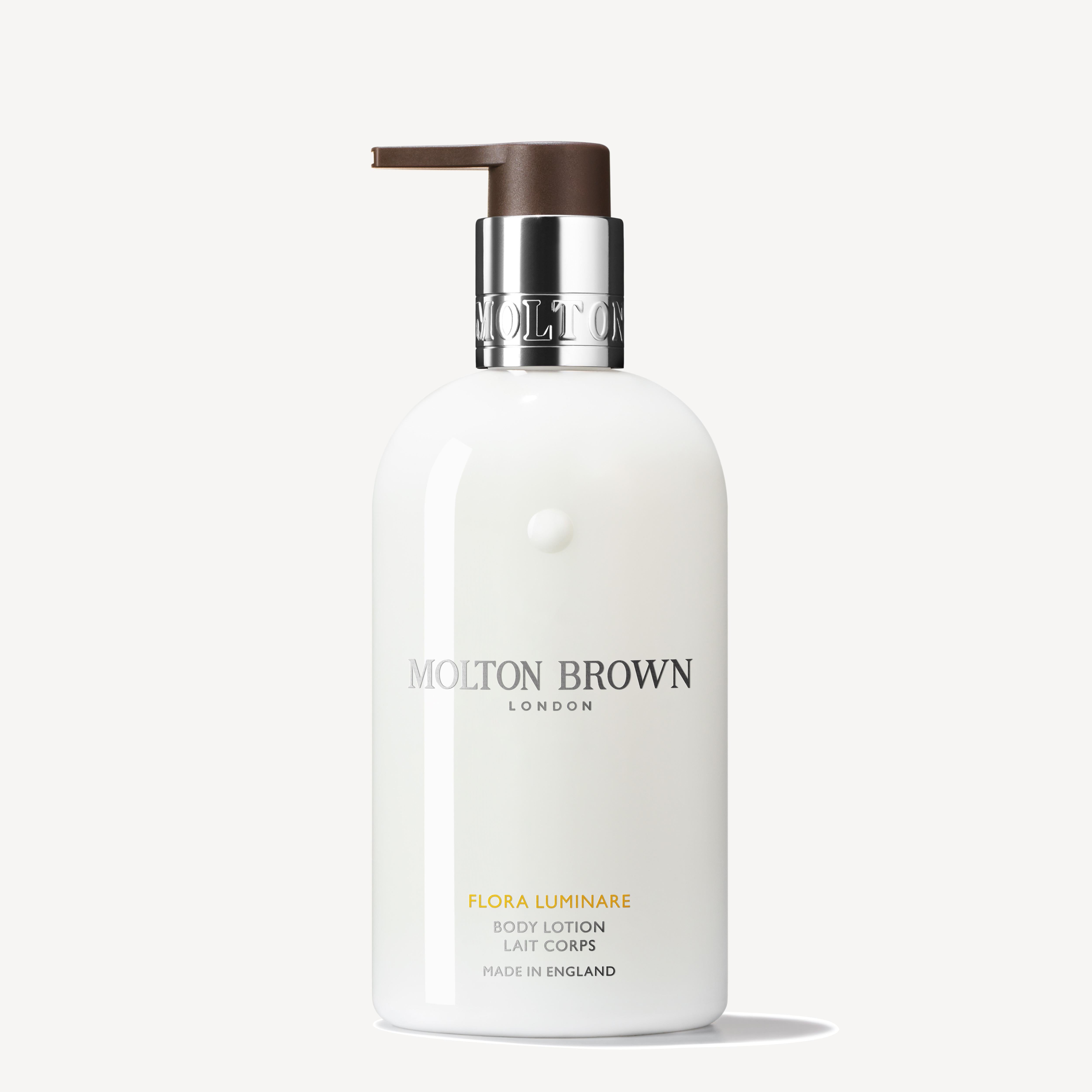 The Flora Luminare Body Lotion 300ml from MoltonBrown Cyprus features a white bottle with a pump dispenser, elegantly displaying the brand's label and logo adorned with silver and brown accents. This fragranced moisturizer, crafted in England, adds a luxurious touch to your skincare routine.
