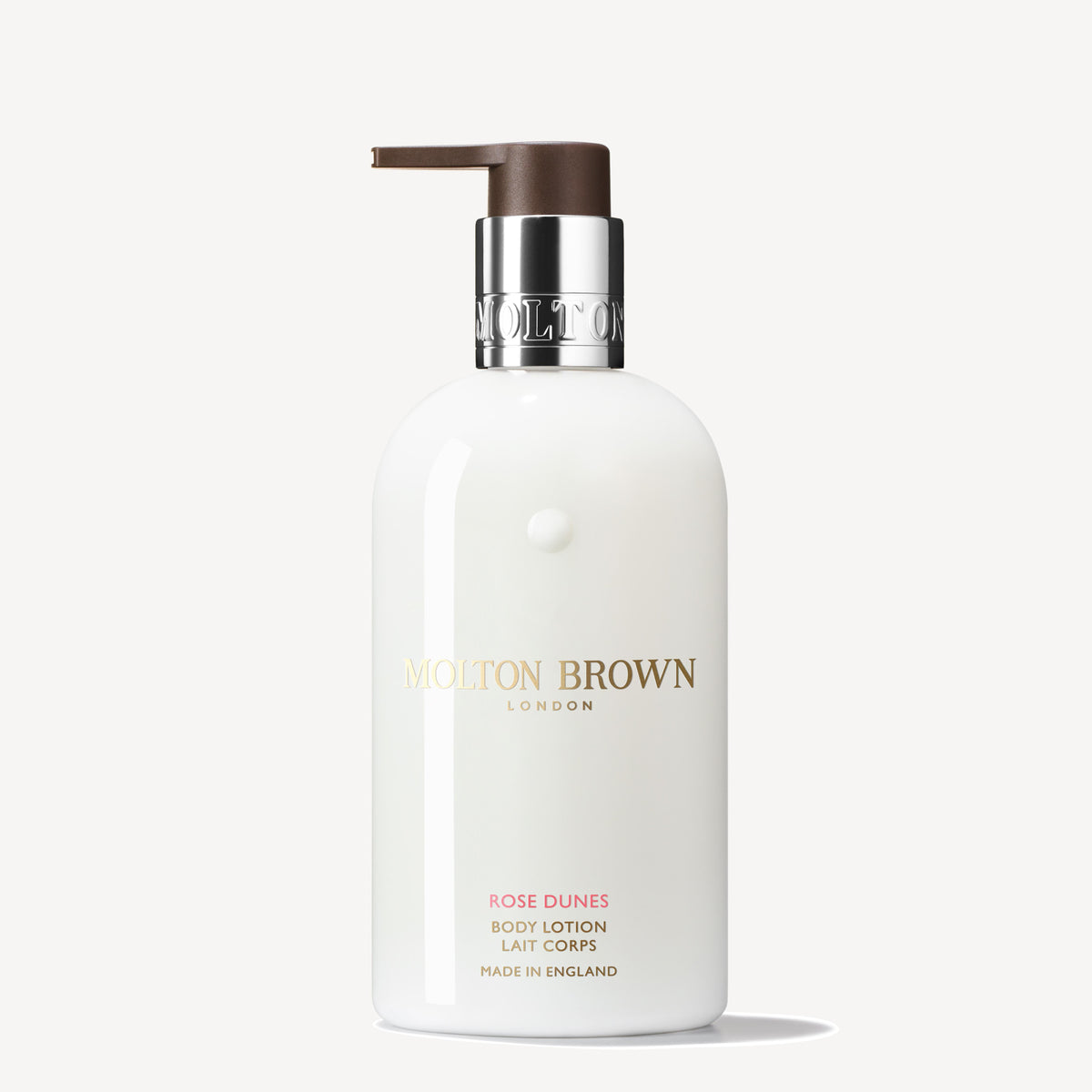 A white bottle of MoltonBrown Cyprus's Rose Dunes Body Lotion 300ml features a gold pump dispenser and offers an ambery-rose scent. The label displays the brand's logo and product details in gold text against a plain off-white background, enhancing its elegance and promise of hydration.