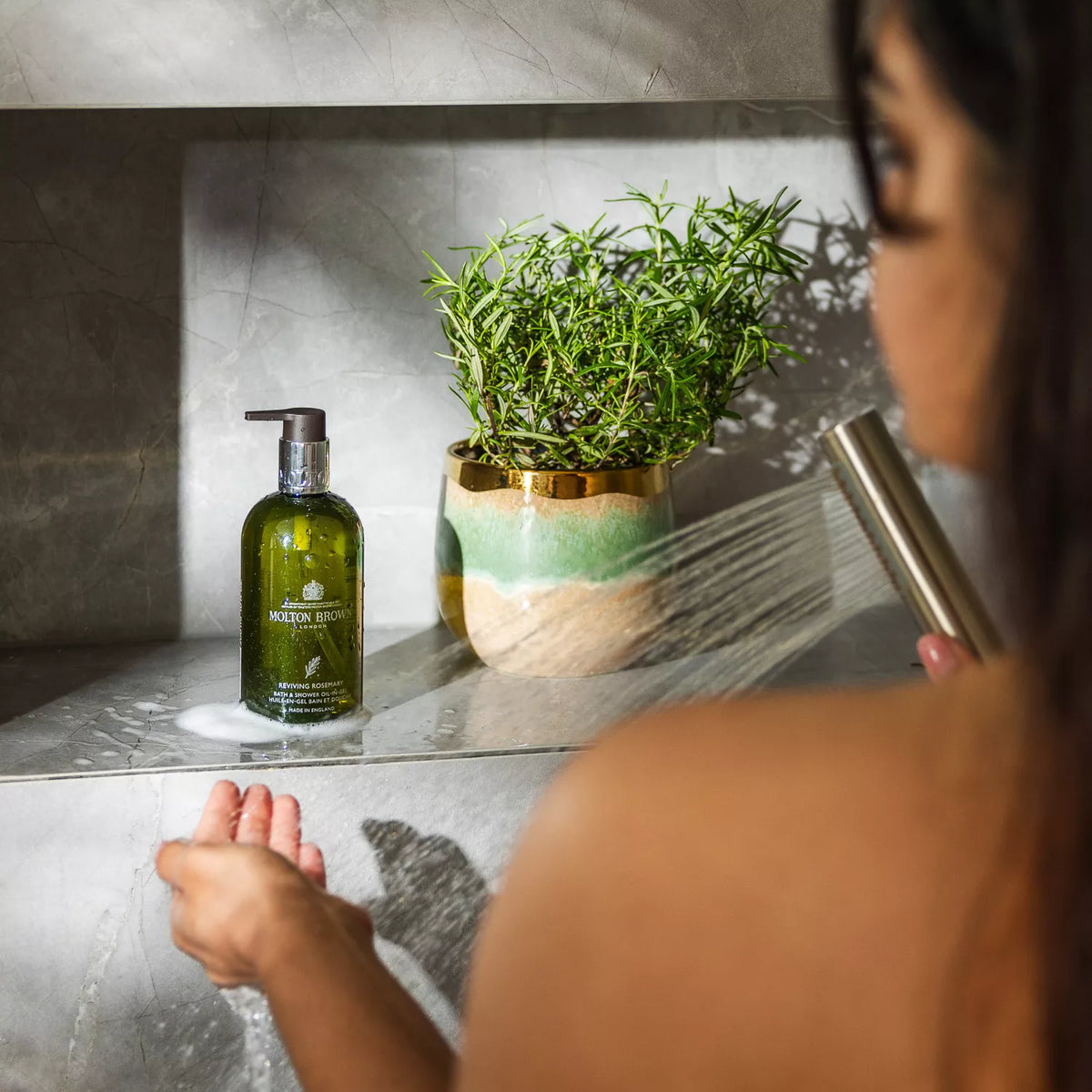 A person showers in a modern bathroom using MoltonBrown Cypruss Reviving Rosemary Bath & Shower Oil-In-Gel, which turns into a luxurious milky lather. A nearby potted plant with green leaves enhances the serene atmosphere.