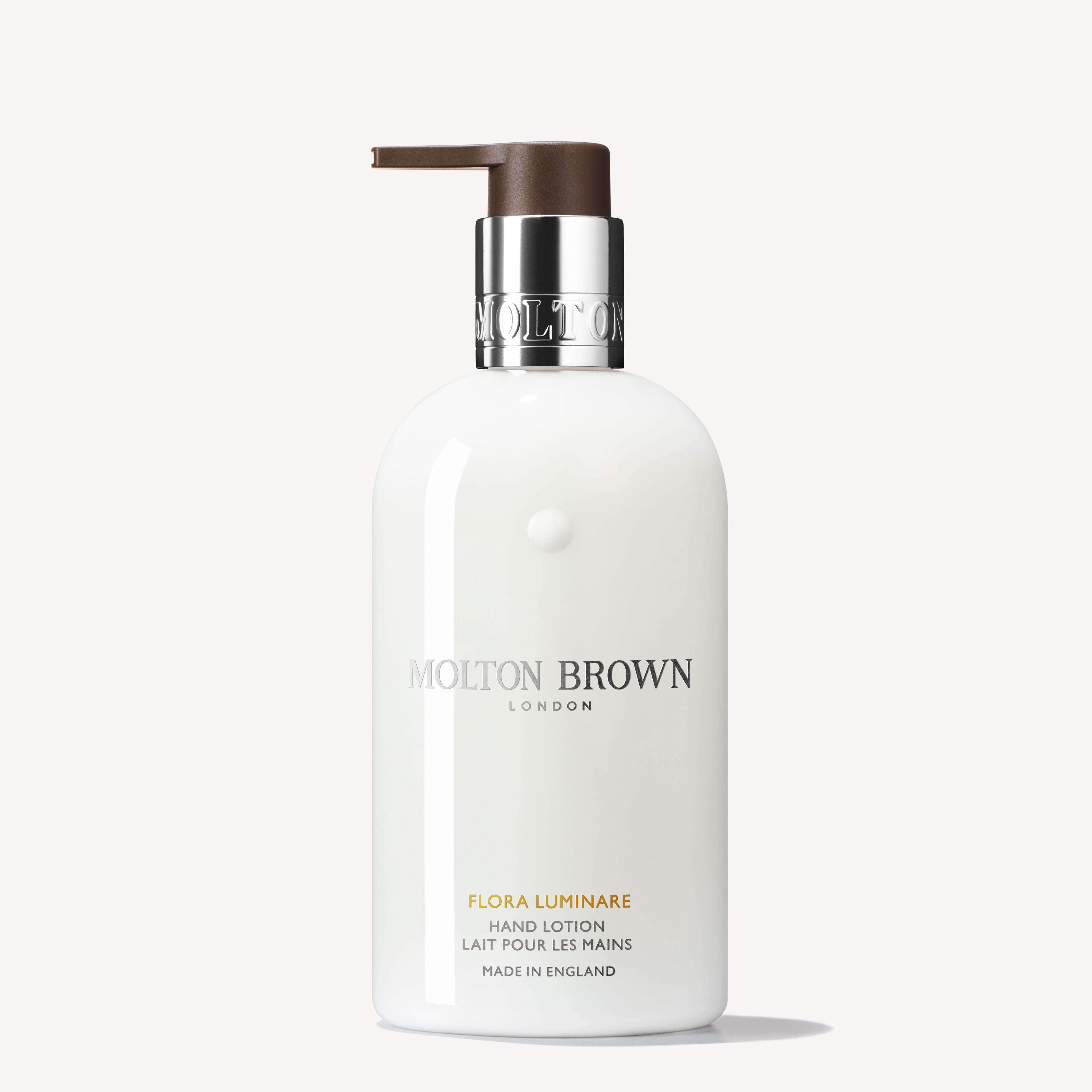 The Flora Luminare Hand Lotion 300ml by MoltonBrown Cyprus comes in a sleek white bottle with a minimalist design, accented by a brown pump top. The label prominently showcases the brand name, product type, and notes its English origin. Experience the indulgent fragrance of white florals it provides.