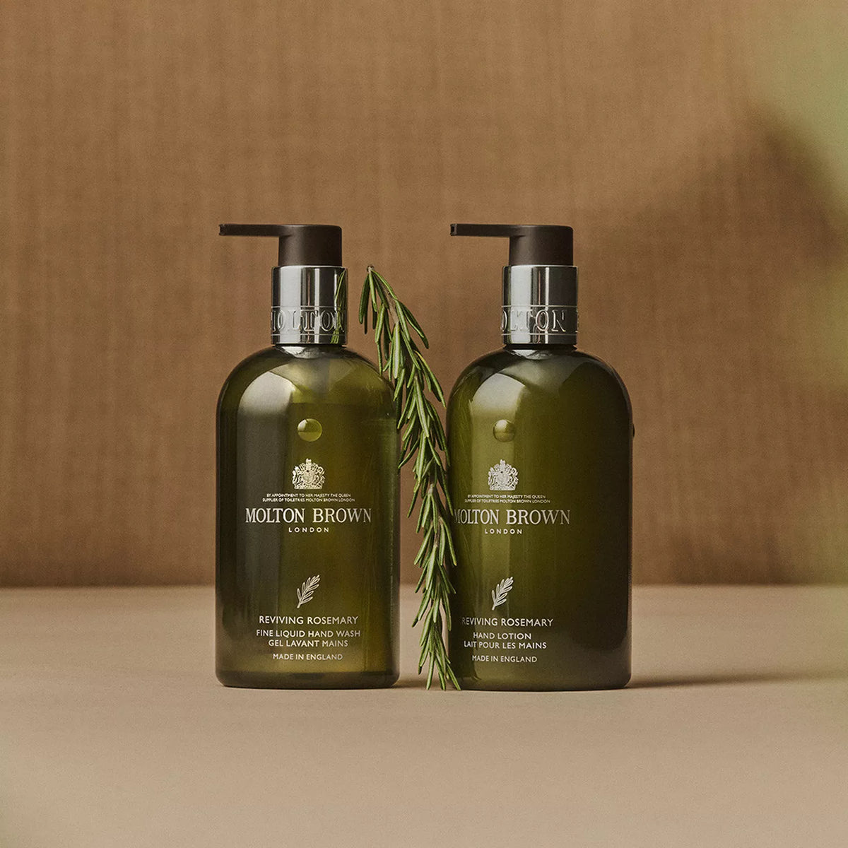 Two green MoltonBrown Cyprus Reviving Rosemary Hand Lotion 300ml bottles with pump dispensers stand side by side on a beige surface, paired with a rosemary sprig. The background is beige.