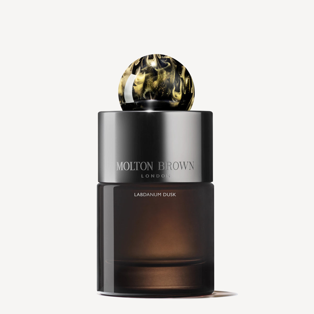 A bottle of Labdanum Dusk Eau de Parfum 100ml by MoltonBrown Cyprus features a dark, glossy design with a decorative marbled spherical cap. It captures an amber woody essence with its rare oudh scent, offering a luxurious fragrance experience against the off-white background.