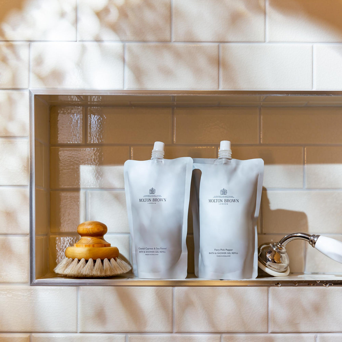 The Coastal Cypress & Sea Fennel Bath & Shower Gel Refill from MoltonBrown Cyprus, with its invigorating marine fragrance, is elegantly showcased in a shower nook adorned with white tiles. Close by, a brush with a wooden handle and a chrome showerhead rest gracefully as gentle sunlight casts shadows on the wallβemphasizing an eco-friendly addition to your bath routine.