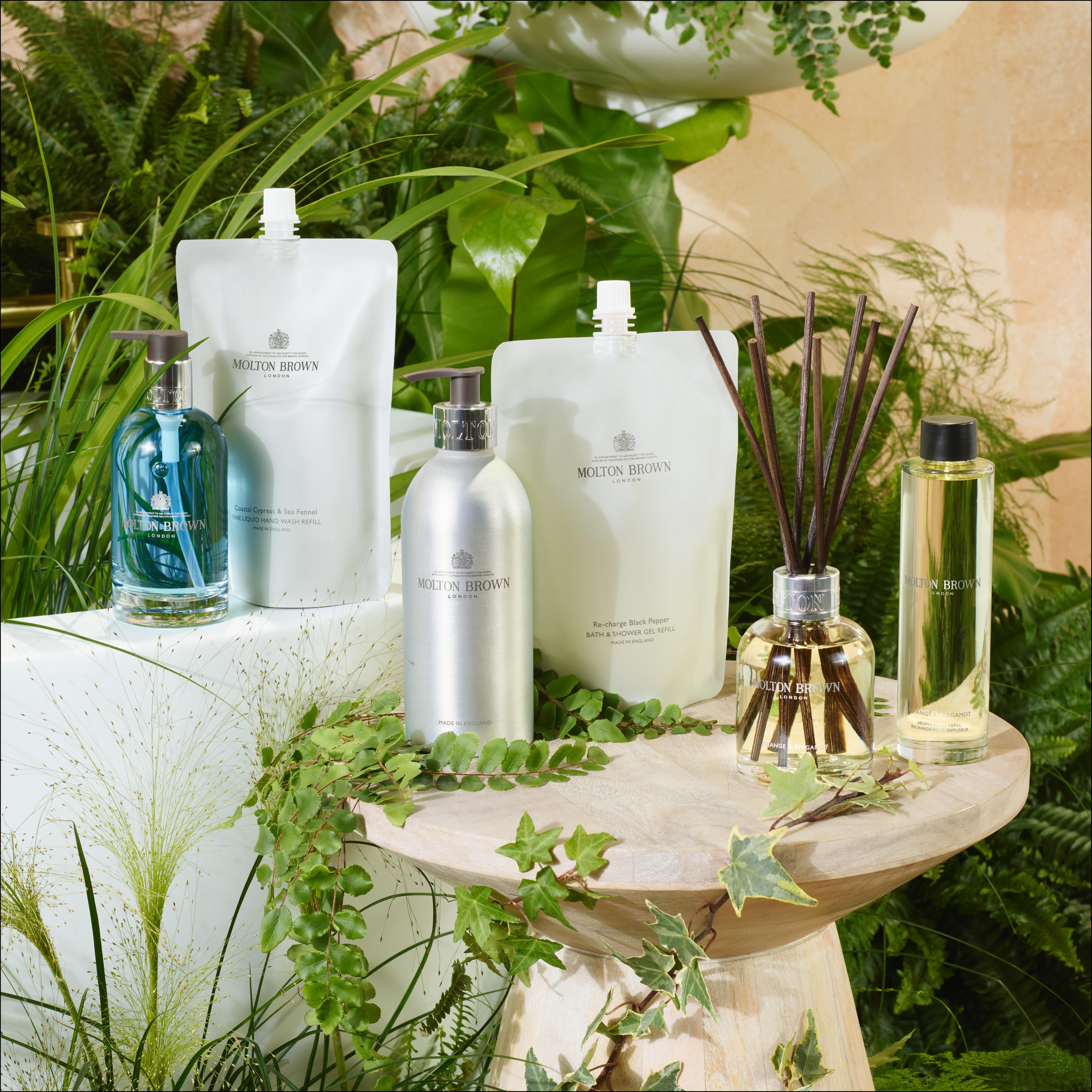 A selection of MoltonBrown Cyprus products, featuring the Re-charge Black Pepper Infinite Bottle 400ml with its eco-friendly refill pouches and a woody-citrus body wash, is arranged on a wooden stool. The display is enhanced by lush green plants, fostering a natural and invigorating ambiance.