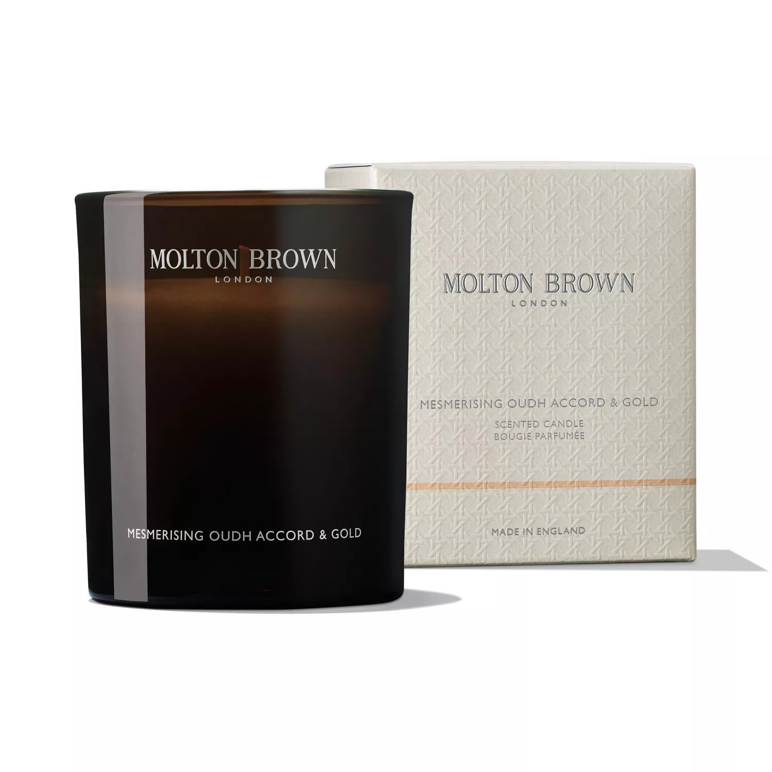 A dark brown luxurious scented candle in a glass holder labeled Mesmerising Oudh Accord & Gold Signature Candle 190g from MoltonBrown Cyprus sits elegantly before its matching cream box, adorned with the same branding and details.