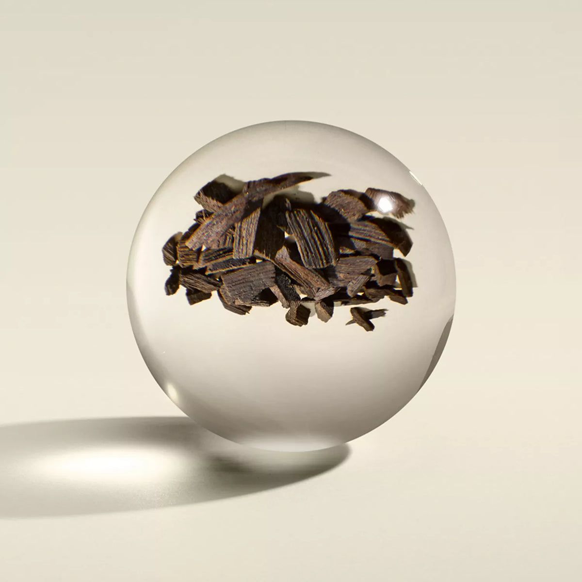 The MoltonBrown Cyprus Mesmerising Oudh Accord & Gold Signature Candle 190g features an opulent clear sphere enclosing jagged dark pieces resembling dried wood chips, evocative of oudh, casting a soft shadow on a light beige surface with the contents appearing suspended inside.