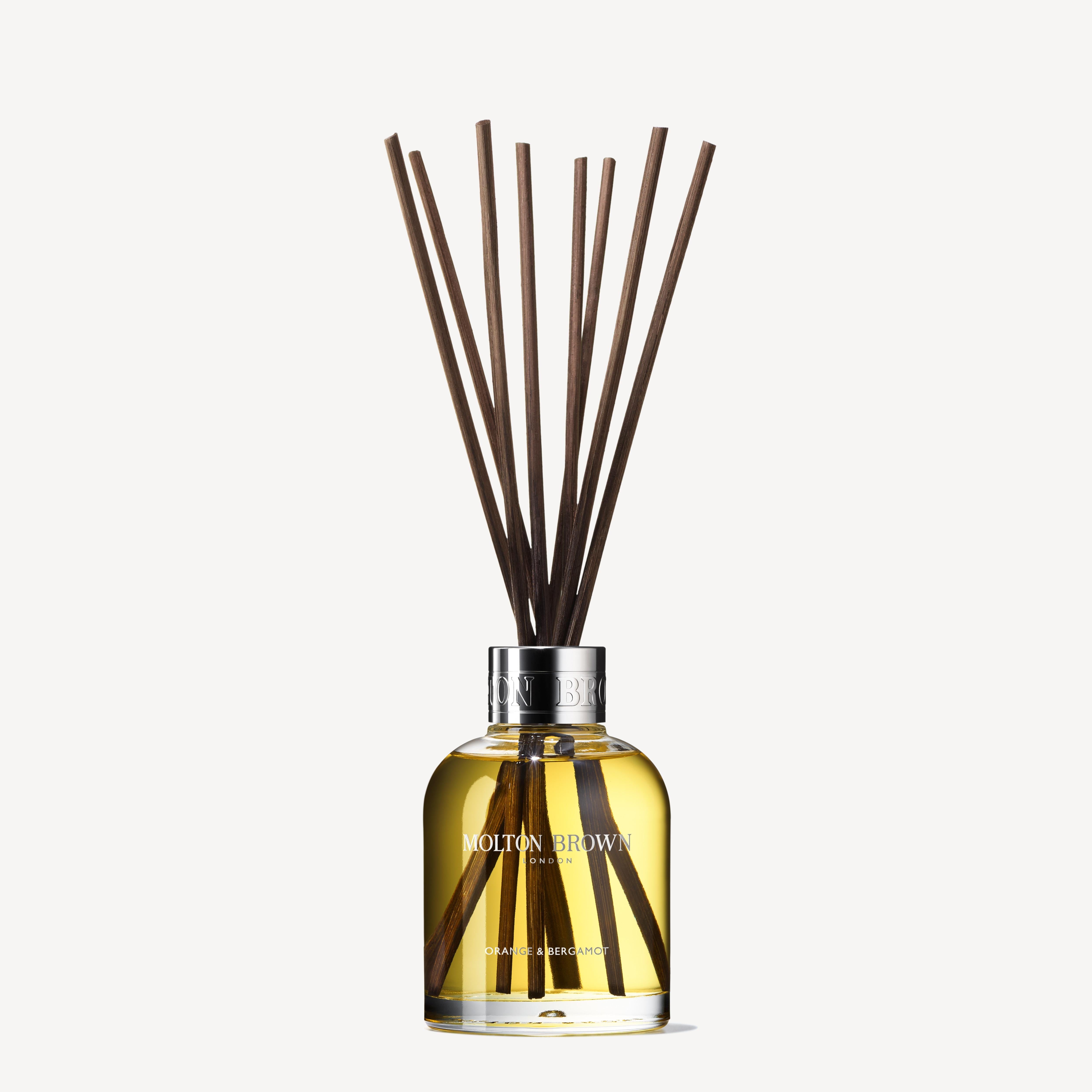 A glass bottle filled with a golden liquid, labeled MoltonBrown Cyprus, features dark reed sticks protruding from the top and serves as the Orange & Bergamot Aroma Reeds 150ml diffuser. The light off-white backdrop accentuates its elegance.