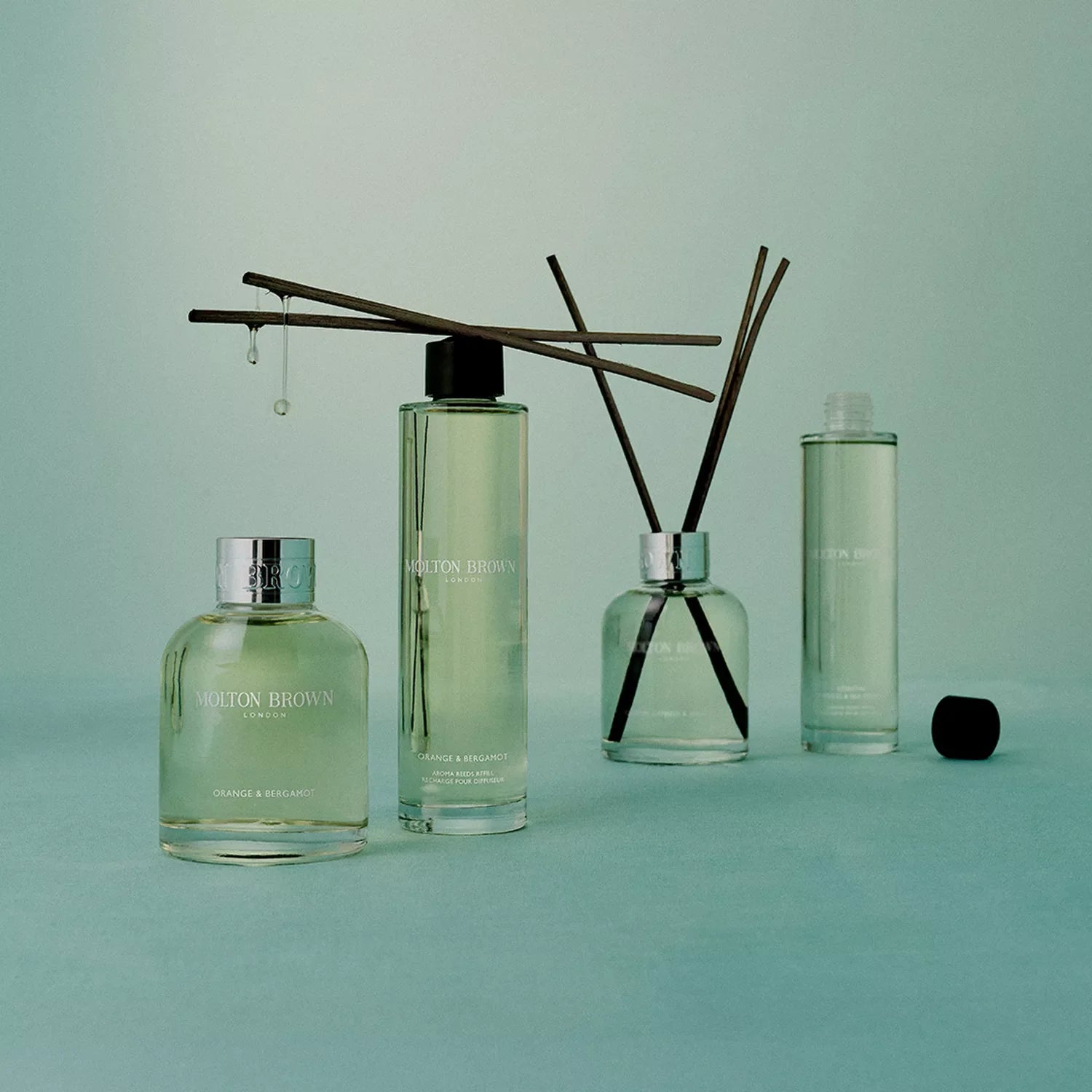 Four bottles of MoltonBrown Cyprus Mesmerising Oudh Accord & Gold Aroma Reeds Refill 150ml are shown on a light green surface; two hold aroma reeds, and one features a dropper with liquid dripping from it.