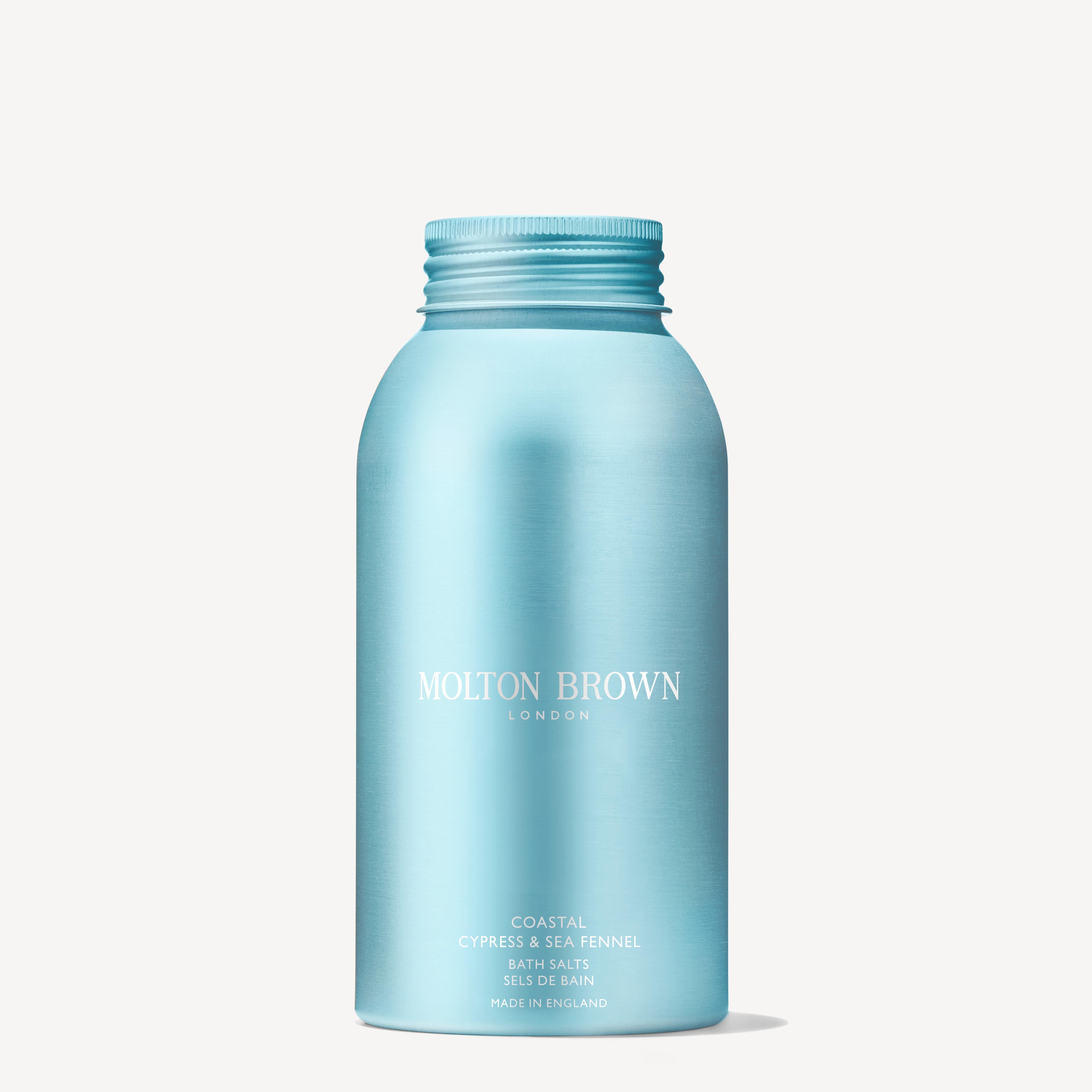 A hand holds a blue bottle of MoltonBrown Cyprus Coastal Cypress & Sea Fennel Bath Salts 300g above a partially filled bathtub with running water. The calming aroma of Coastal Cypress harmonizes beautifully with the bathroom's sleek chrome faucet fixtures, creating an inviting escape.
