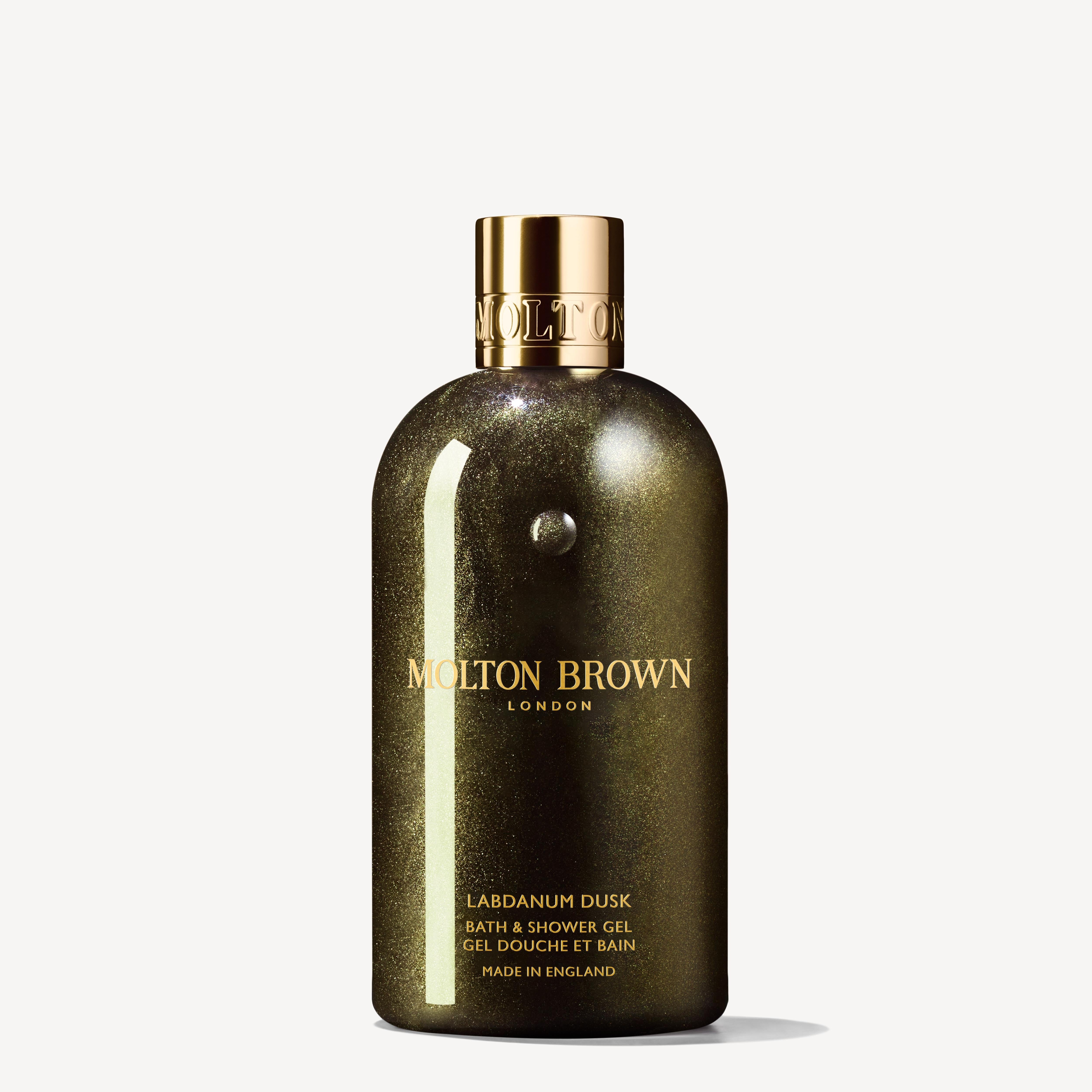 The image showcases a Labdanum Dusk Bath & Shower Gel 300ml bottle from MoltonBrown Cyprus, featuring its glittery dark green packaging accented by a gold cap and lettering. This luxurious presentation invites you to indulge in an ambery-woody fragrance with hints of opulent oudh. Proudly displayed is the note "Made in England.