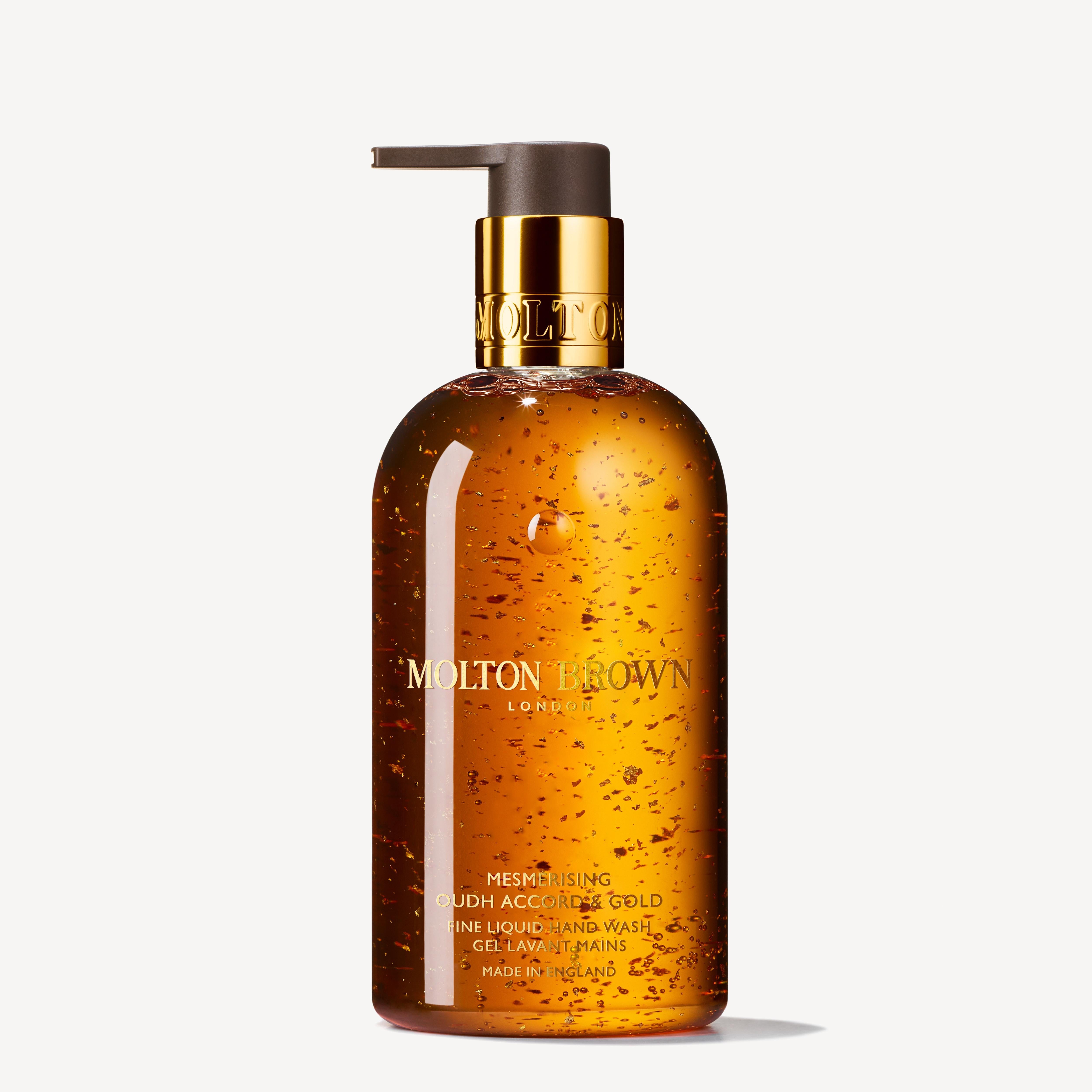 A 300ml bottle of MoltonBrown Cyprus Mesmerising Oudh Accord & Gold Fine Liquid Hand Wash, equipped with a pump dispenser. The elegant golden-brown design showcases 23.5-carat golden flakes shimmering inside, with the label prominently displaying the brand name and product title.