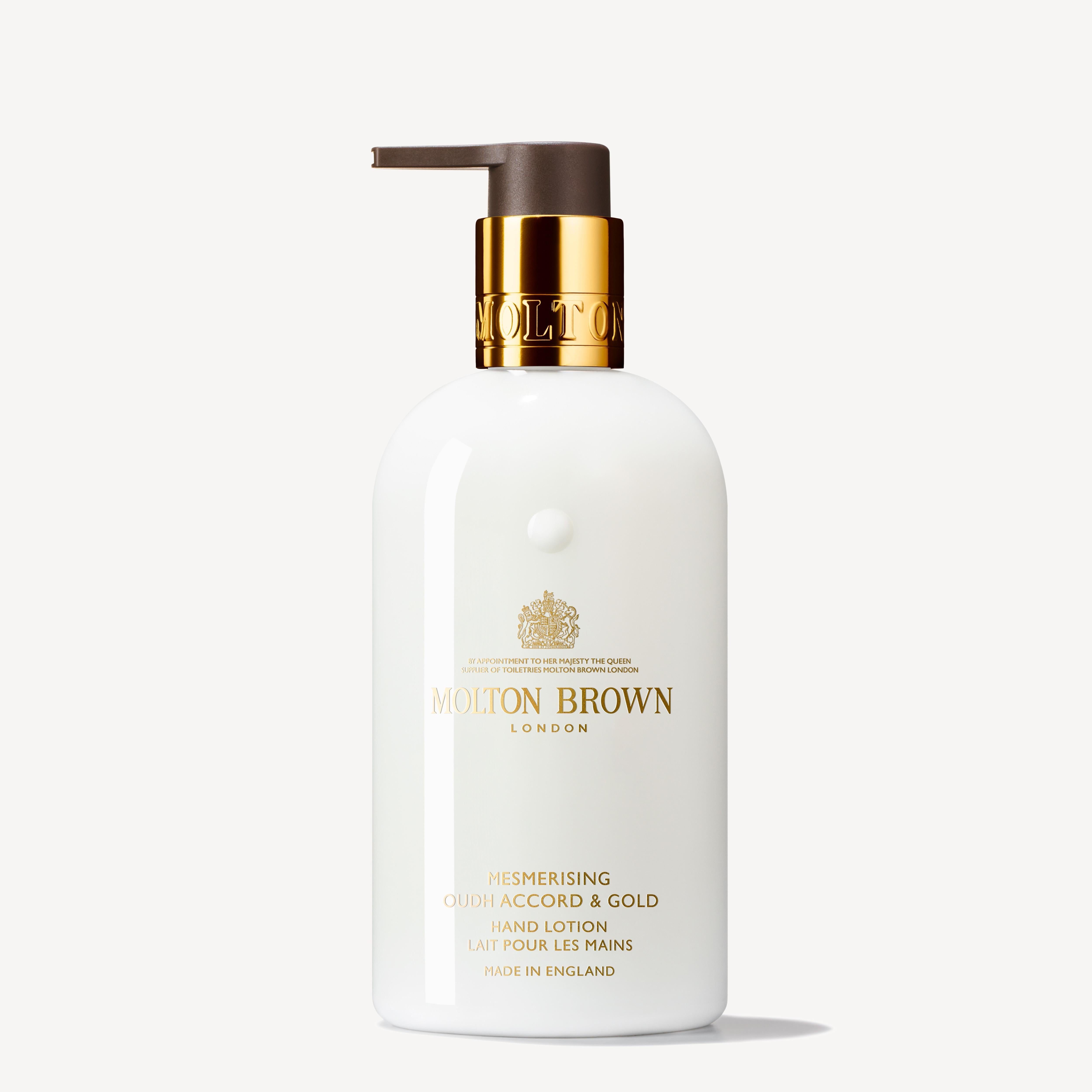 A white bottle of MoltonBrown Cyprus' luxe Mesmerising Oudh Accord & Gold Hand Lotion, featuring a gold pump. The design is elegantly completed with gold and black text, along with an emblem above the brand name. The off-white background accentuates the allure of this luxurious vegan formula infused with honey.