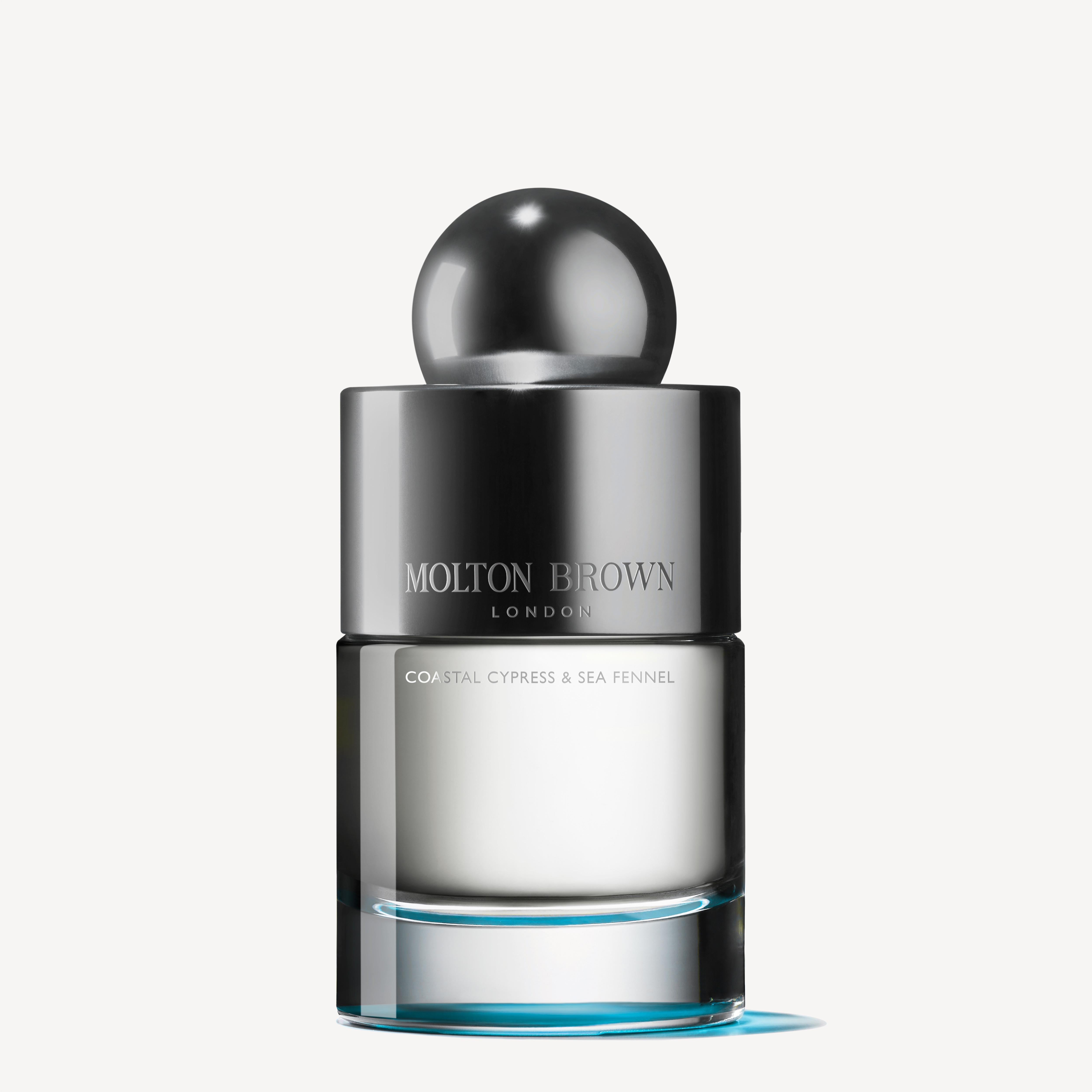A bottle of Coastal Cypress & Sea Fennel Eau de Toilette 100ml by MoltonBrown Cyprus. The cylindrical design is highlighted by a metallic spherical cap, with a clear section displaying the pale liquid. Accentuated with hints of fig leaves and cedarwood, the sleek label retains its minimalist charm.